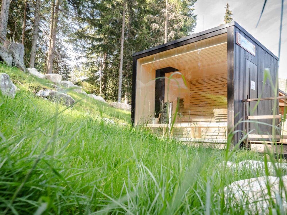 This,Is,The,Way,I,Like,To,Take,Sauna Cube-Sauna mit großer Glasfront und Holzverkleidung, steht auf Holzterrasse mit Panoramablick aus dem Garten.