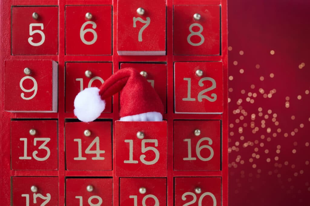 Roter Adventskalender aus Holz mit goldfarbenen Zahlen, Tür 15 geöffnet, darin roter Weihnachtsmannmütze mit weißem Bommel.