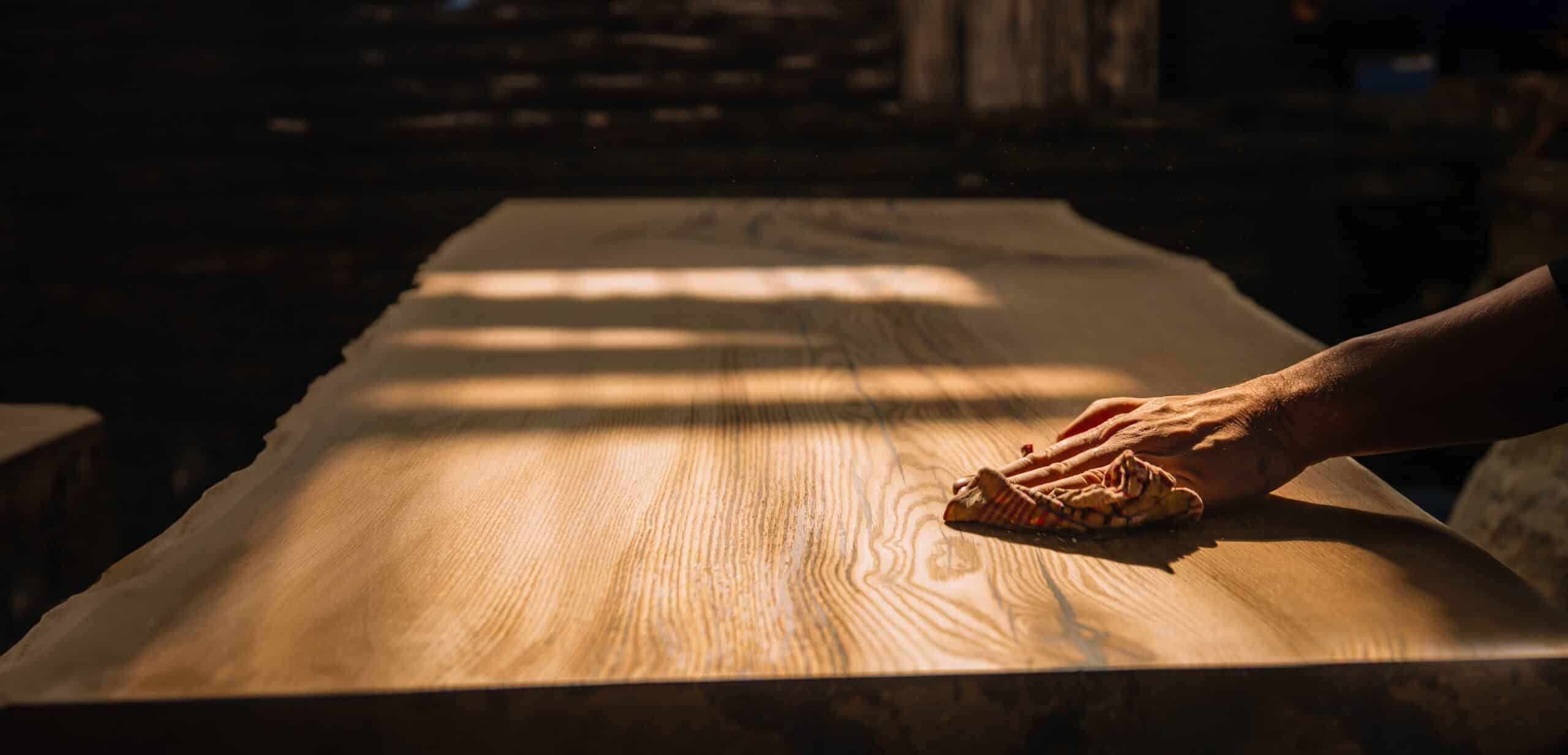 Hand wischt mit Tuch über eine helle Holzoberfläche eines Tisches, Sonnenlicht fällt schräg darauf.