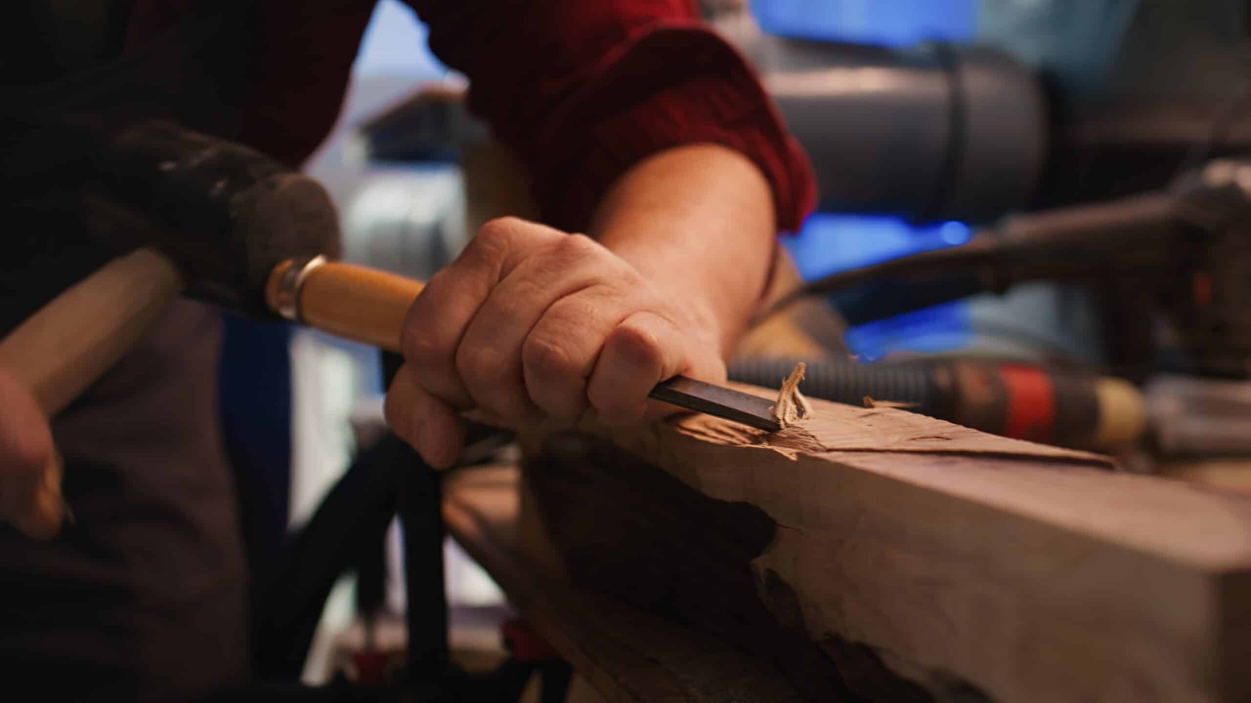 Hand hält Holzbeitel und schnitzt an einem Stück Holz in einer Werkstatt mit unscharfem Hintergrund.