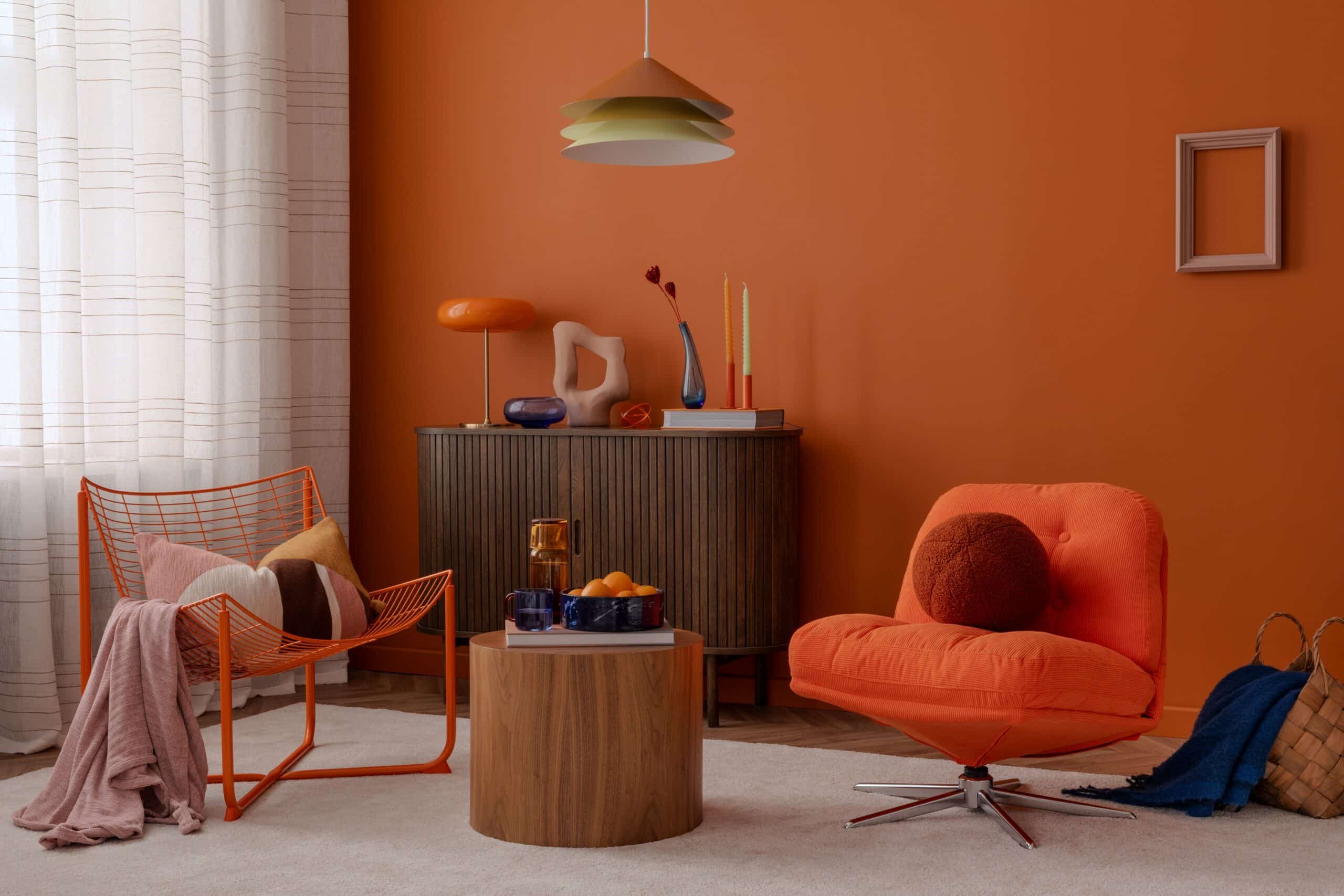 Oranger Sessel mit rundem Kissen, daneben oranger Drahtstuhl mit Kissen und Decke, mittig runder Holztisch, im Hintergrund Sideboard mit Vasen und Kerzen vor oranger Wand.