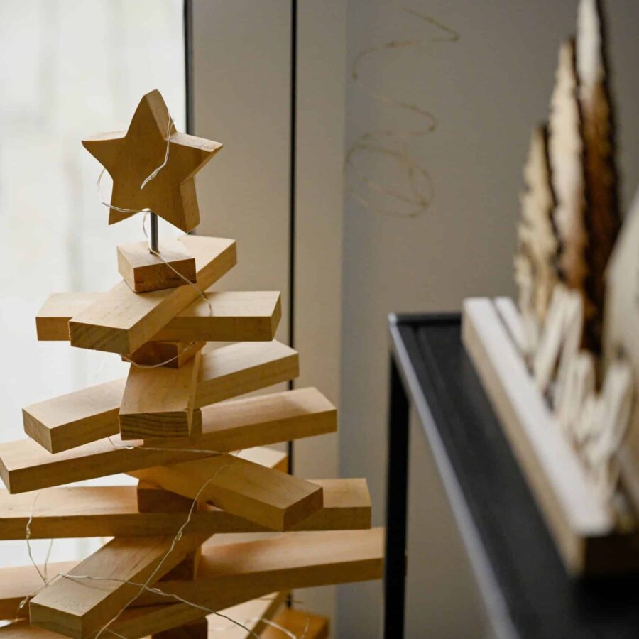 Holz-Weihnachtsbaum mit gestapelten, rechteckigen Stäben und einem Stern an der Spitze, unscharfe Deko im Hintergrund.