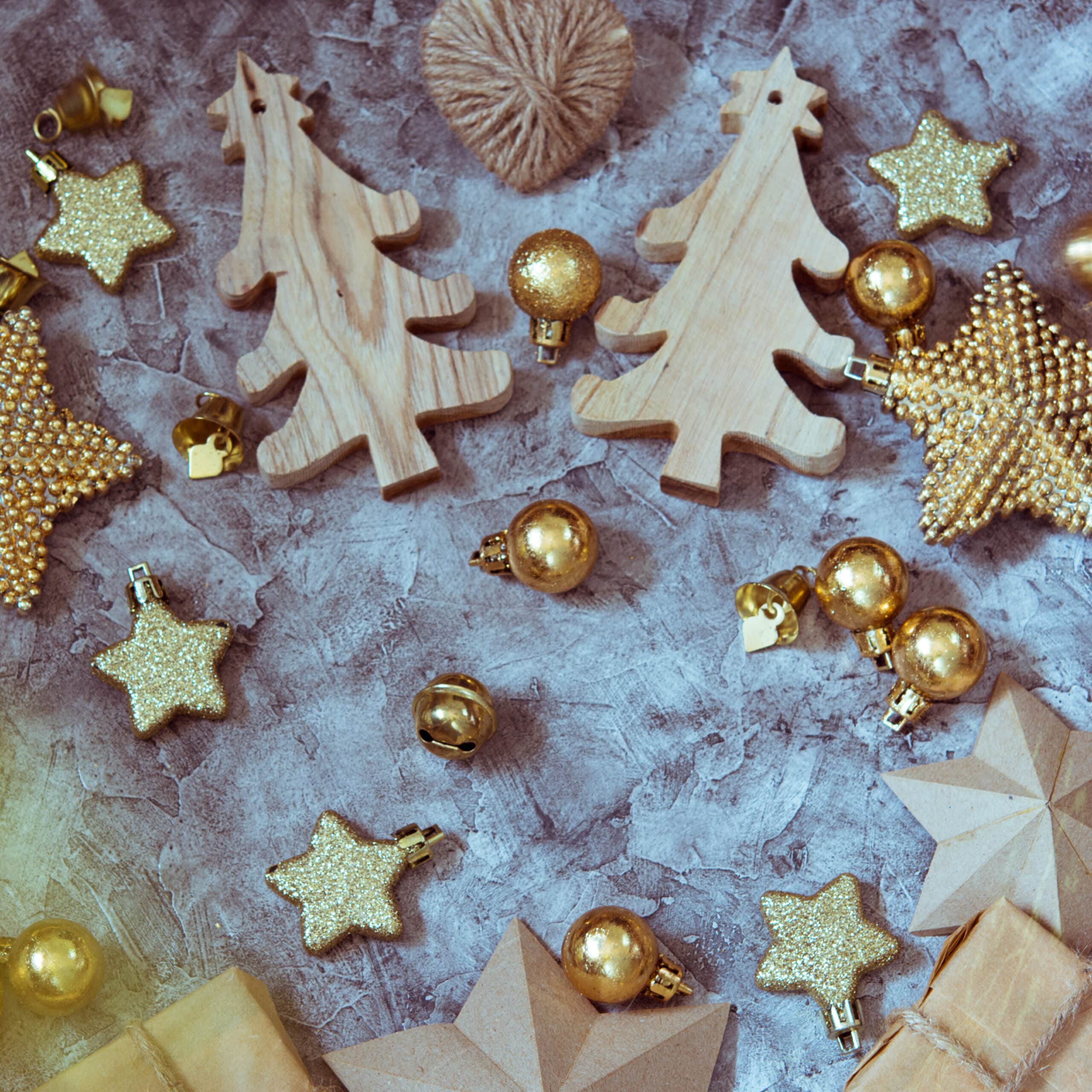 Zwei hölzerne Weihnachtsbäume, mehrere goldene Glitzersterne, goldene Kugeln und ein Wollknäuel auf grauem Untergrund.