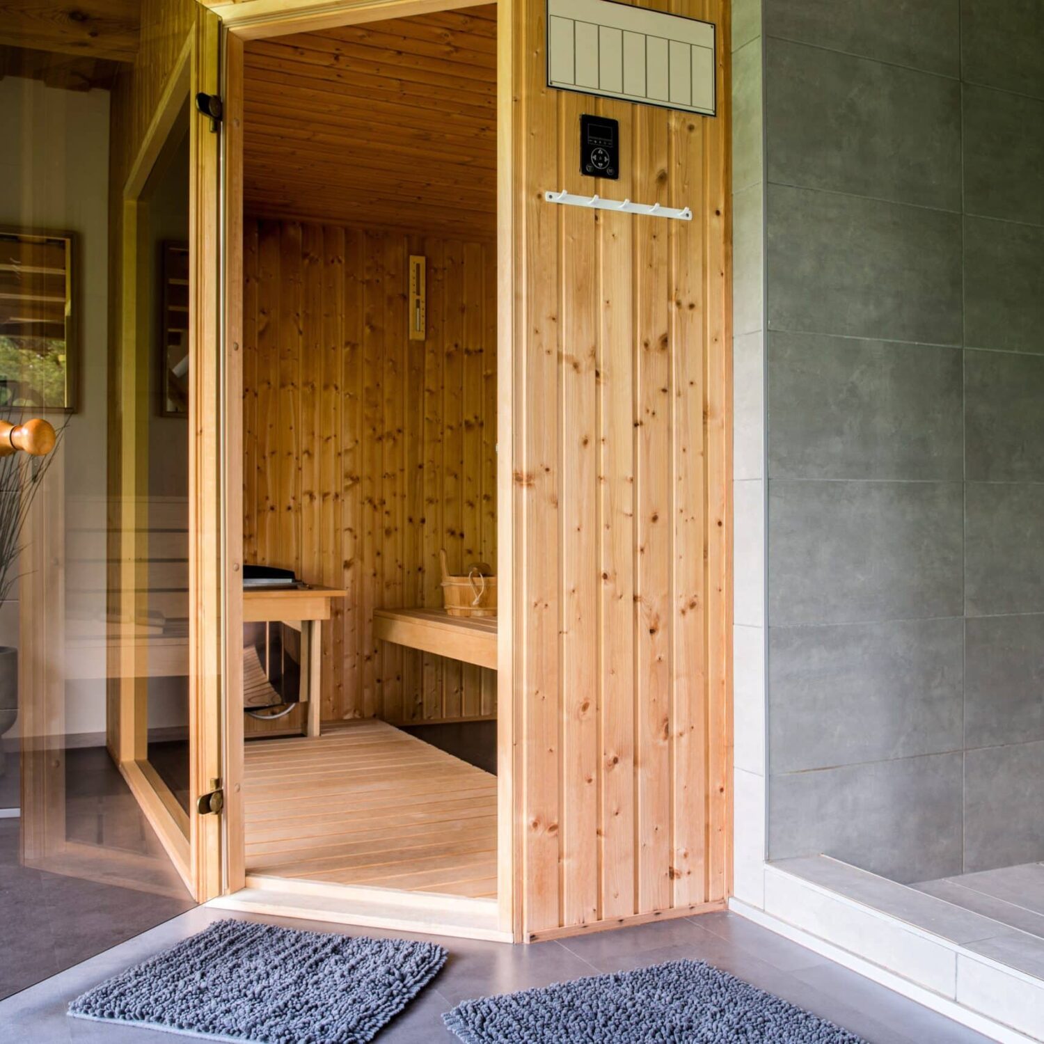 Wooden,Sauna,With,Benches.,The,Sauna,Is,Empty.,The,Wooden Offene Sauna mit Holzwänden und Glasfront, davor zwei graue Fußmatten auf grauem Boden, rechts graue Fliesenwand mit Duschbereich.