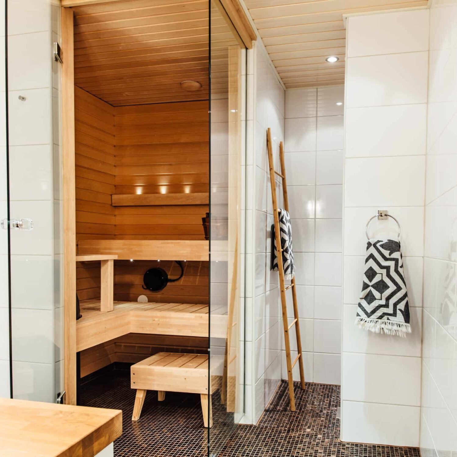 Finnish,Bathroom,With,A,Small,Wooden,Sauna,And,Mosaic,Tile Badezimmer mit moderner Sauna aus hellem Holz, Glastür, zwei Handtüchern mit geometrischem Muster und dunklem Mosaikfliesenboden.