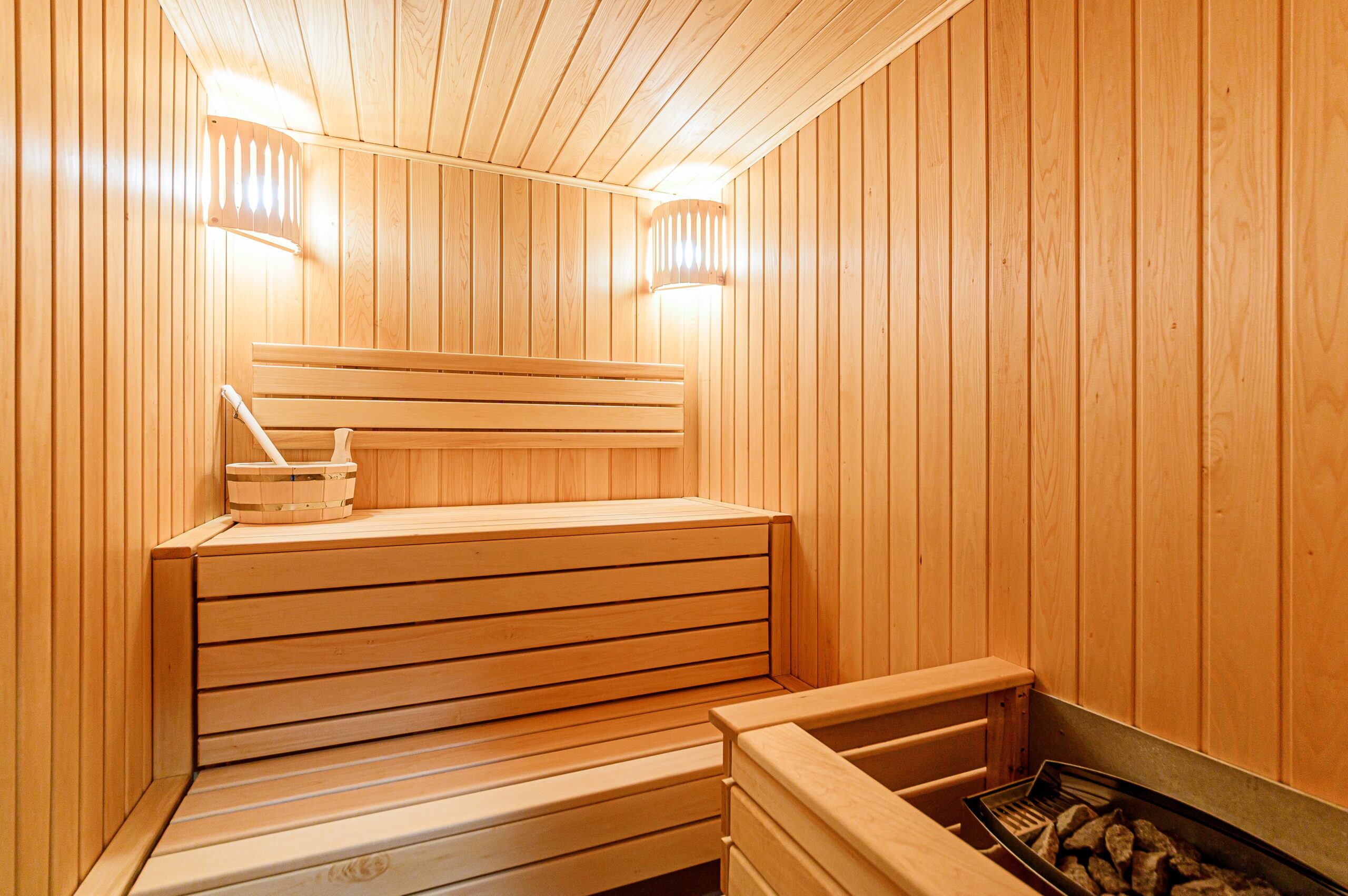Cozy,Apartment,Interior,Features,A,Wooden,Sauna,,Blending,Modern,Living Innenraum einer Sauna mit Holzbänken, Holzwänden, zwei Wandlampen und einem Saunaofen mit Steinen.