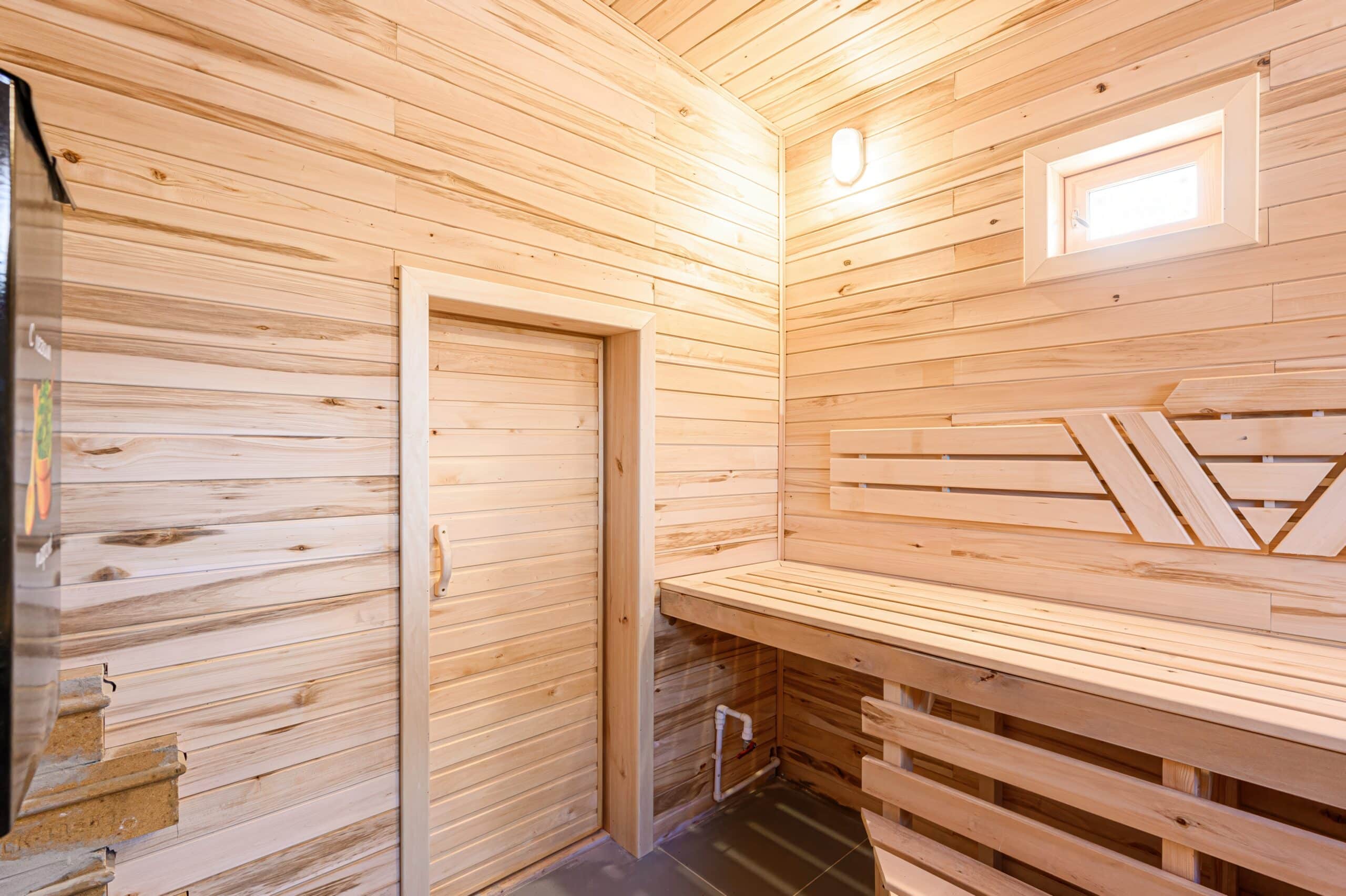 Sauna,With,Wooden,Walls,And,Benches,,A,Door,,A,Small Innensauna mit hellen Holzpaneelen, Holztür, zwei Holzbänken in L-Form und kleinem Fenster mit Lichtquelle.