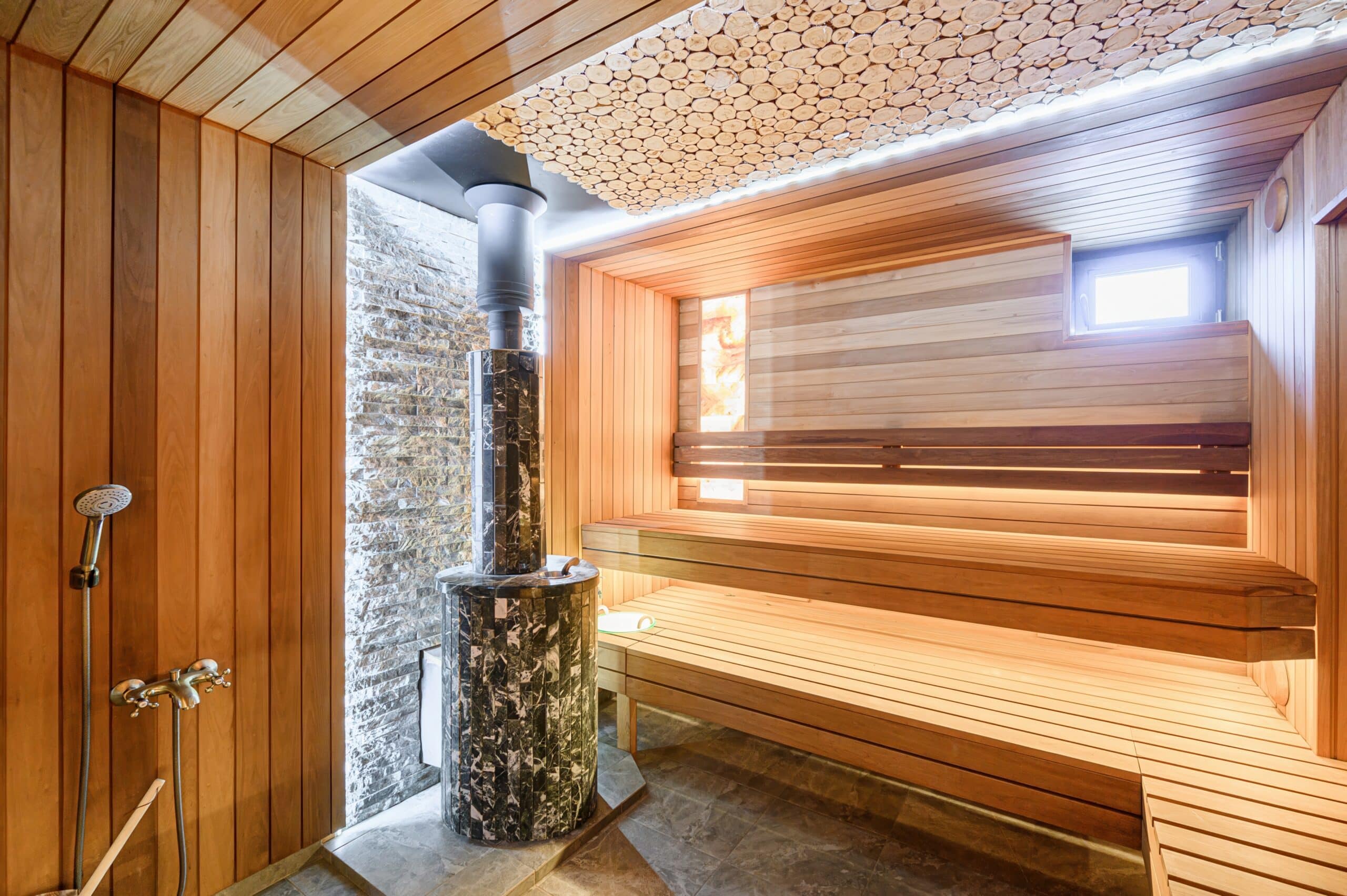 Warm,Sauna,Interior.,Wooden,Benches,,Stone-clad,Stove,,And,Unique,Wooden Innensauna mit Holzverkleidung, zwei Holzbänken, einem runden, gefliesten Saunaofen mit Steinen und einem Fenster an der Wand.