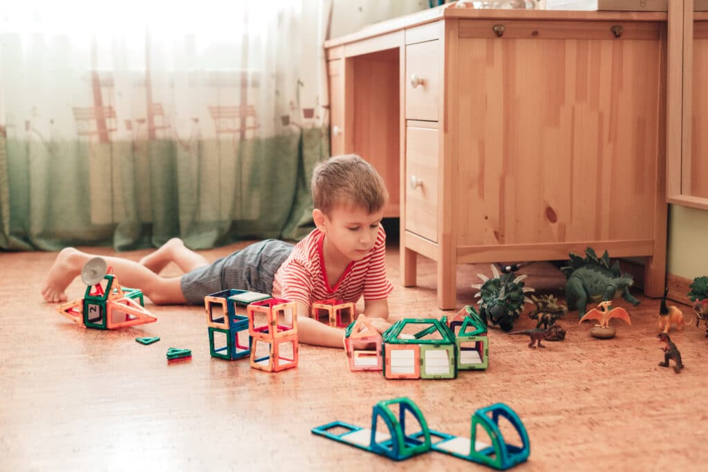 A,Boy,Plays,With,Dinosaurs,And,Magnetic,Constructor,In,A Kind liegt auf einem Korkboden und spielt mit bunten magnetischen Bausteinen und Dinosaurierfiguren neben einem Holzmöbelstück.