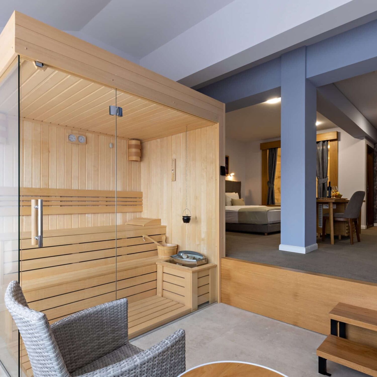 Small,Sauna,In,Hotel,Room Holzsauna mit Glasfront und zwei Sitzbänken, steht in einem hellen Raum neben einem Sessel mit Kissen und einem kleinen Tisch mit Handtüchern, im Hintergrund steht ein Bett.