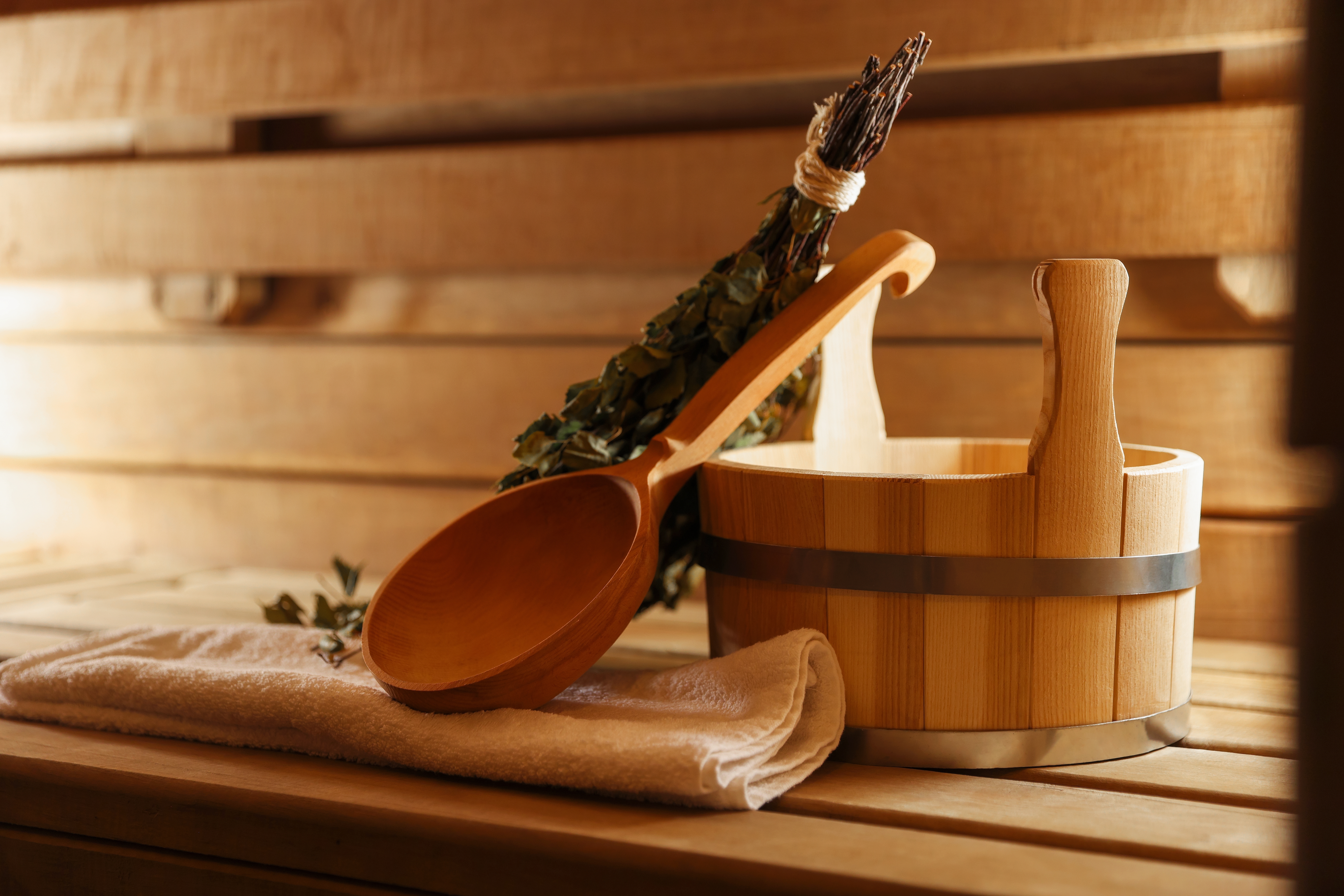 Ladle,,Towel,,Bucket,And,Whisk,On,Wooden,Bench,In,Sauna Saunaeimer aus Holz mit Holzlöffel neben einem Handtuch und einem Bund Kräuter in einer Sauna.
