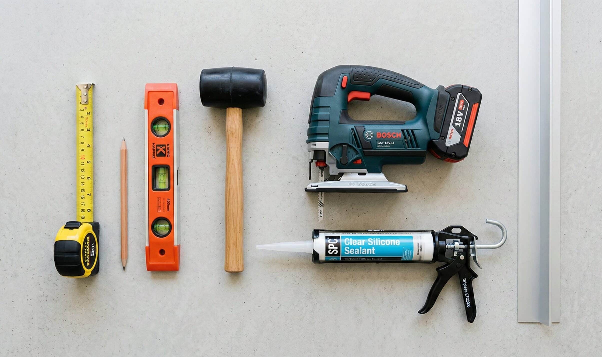 werkzeug_paneele Werkzeug und Materialien zum Heimwerken auf hellem Untergrund angeordnet, darunter Maßband, Bleistift, Wasserwaage, Gummihammer, Akku-Stichsäge, Kartuschenpistole mit Silikondichtstoff und Aluminiumprofil